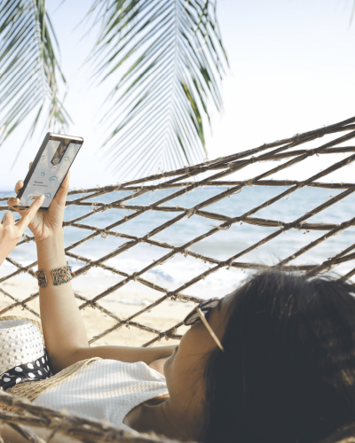 Une personne est en vacances sur un hamac et est tranquille d'esprit grâce à l'application mobile OtioHome qu'elle consulte depuis la plage pour veiller sur sa maison